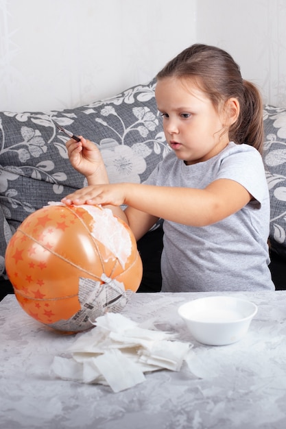 Un niño se sienta en el sofá de la sala de estar y hace una calabaza de papel maché para Halloween con un globo.