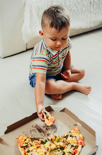 El niño se sienta en el suelo y come pizza muy apetecible y golosa, en pantalón corto y camiseta