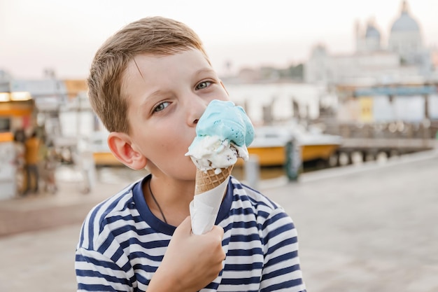 Un niño de siete años con una camiseta a rayas se sienta en el paseo marítimo de Venecia por la noche y come helado Turismo y recreación en Europa con niños