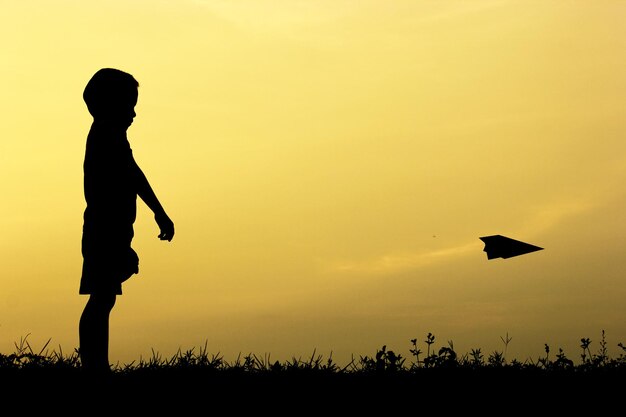 Foto niño silueta jugando con un avión de papel sobre el campo contra el cielo durante la puesta de sol