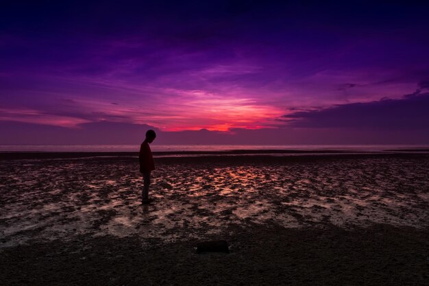 Foto niño silueta de pie en la playa contra el cielo durante la puesta de sol