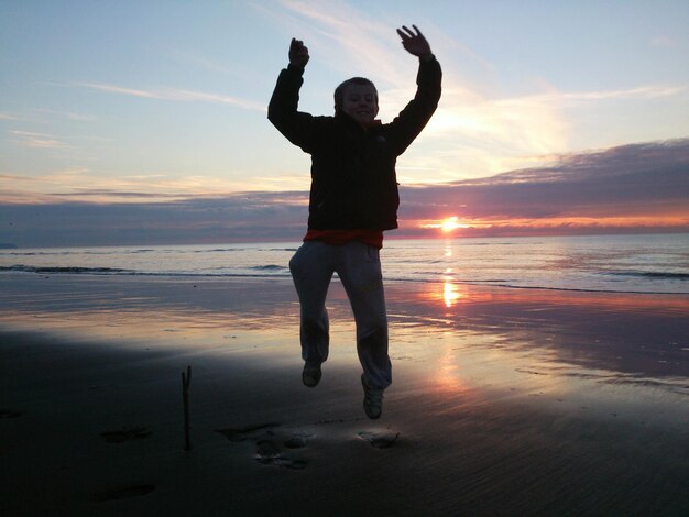 Niño silueta saltando con los brazos levantados en la playa contra el cielo durante la puesta de sol