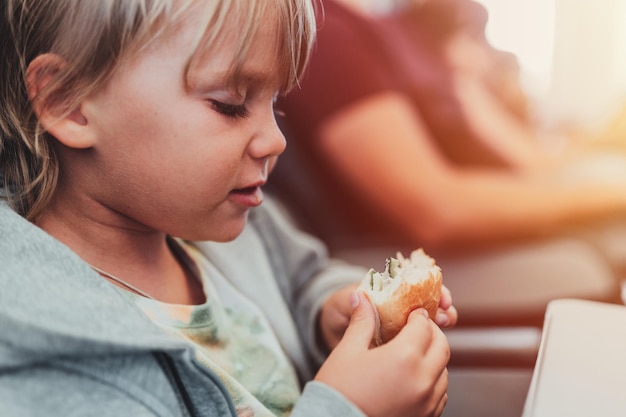 Un niño sincero de cinco años come hamburguesas o sándwiches sentado en el asiento del avión en un vuelo que viaja desde el aeropuerto. Los niños toman un bocado.