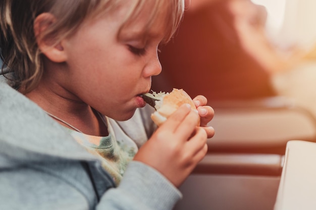 Un niño sincero de cinco años come hamburguesas o sándwiches sentado en el asiento del avión en un vuelo que viaja desde el aeropuerto. Los niños toman un bocado.