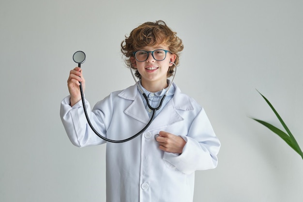 Niño sincero con gafas y bata médica con estetoscopio mirando a la cámara durante el día