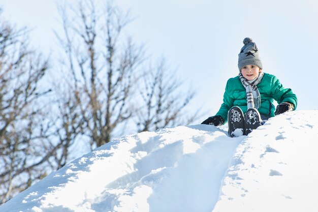 El niño con sombrero, bufanda y chaqueta verde se desliza hacia abajo desde un tobogán de nieve en la espalda.