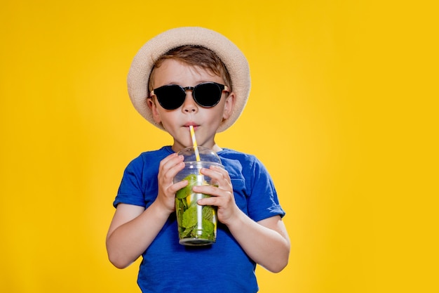 Niño con sombrero de paja y camiseta azul bebe mojito en vaso de plástico sobre fondo amarillo