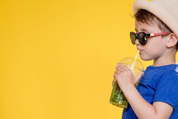 Niño con sombrero de paja y camiseta azul bebe mojito en vaso de plástico sobre fondo amarillo