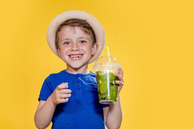 Niño con sombrero de paja y camiseta azul tiene mojito en vaso de plástico sobre fondo amarillo