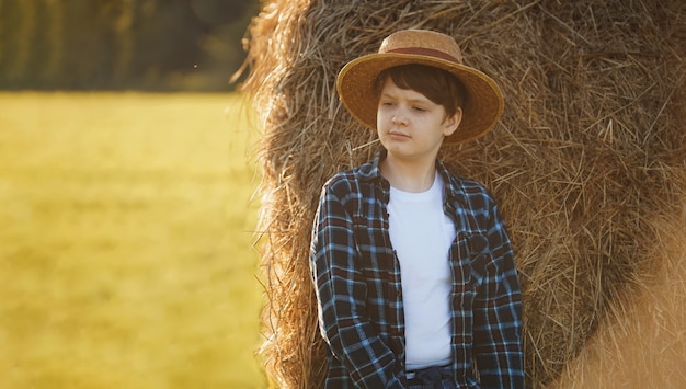 Niño con sombrero de paja de pie cerca de pacas de heno