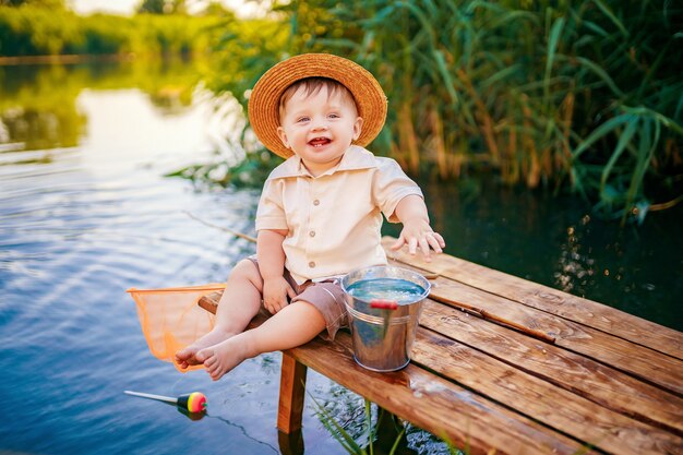 Niño con sombrero de paja sentado en el borde de un muelle de madera y pescando en el lago al atardecer.