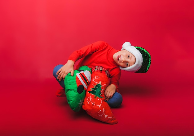 Un niño con un sombrero de Papá Noel está sentado en el suelo con un pequeño árbol de Navidad y regalos de Año Nuevo en un fondo rojo con un espacio para escribir el concepto de Navidad