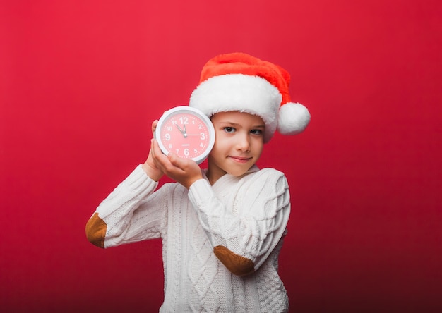 Un niño con un sombrero de Papá Noel sostiene un reloj sobre un fondo de estudio rojo el concepto de Navidad y año nuevo un niño con ropa de punto