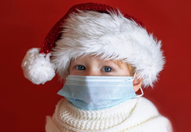 Foto niño en un sombrero rojo de santa está enfermo en navidad, está protegido de virus por una máscara médica