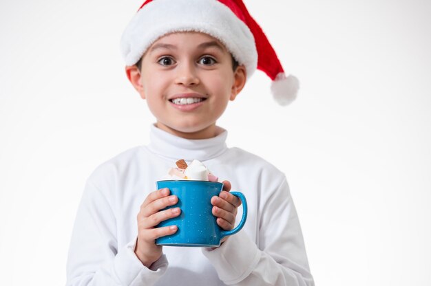 El niño con un sombrero rojo de santa sonríe y sostiene una taza de malvaviscos. Concepto de navidad.