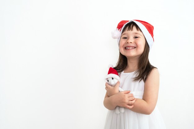 Un niño con sombrero Santa Claus abraza a una liebre tejida blanca. Bebé con gorra de Navidad abraza suavemente un juguete