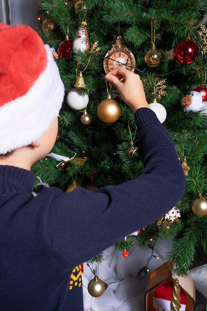 Un niño con un sombrero de Santa Claus cuelga una bola de Navidad en un árbol de Navidad decora la casa para las vacaciones