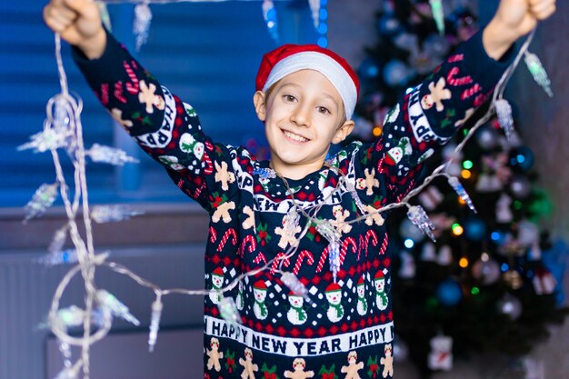 Un niño con un sombrero de Santa Claus con guirnaldas en sus manos en el fondo de un árbol de Navidad con luces de neón