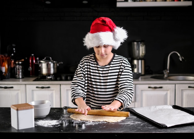 Niño con sombrero de Santa está horneando un pastel de Navidad en la cocina. En tonos negros, rojos y blancos