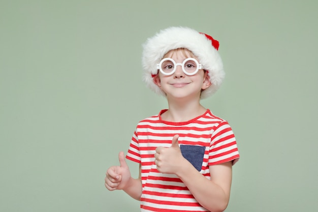 Niño con sombrero de Santa y gafas sonriendo y mostrando los pulgares para arriba. Retrato