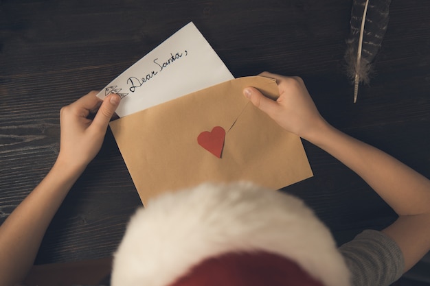 Niño en un sombrero de Santa pone una carta para Santa en un sobre. Vista superior