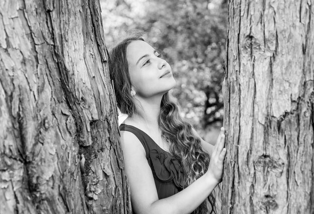 Un niño soñador con cabello largo y ondulado y una mirada de belleza sueña con el tronco de un árbol en el futuro del parque de verano