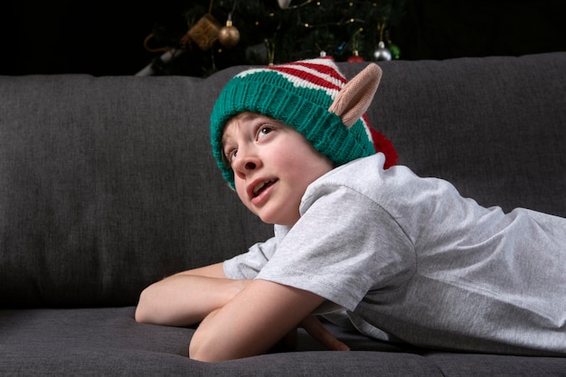 Niño soñador sorprendido con sombrero de elfo de Navidad se encuentra en el sofá en el fondo del árbol de Navidad. Nochebuena