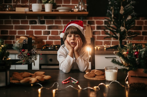 Un niño soñando está sentado en la mesa de la cocina con un suéter blanco y un sombrero rojo de Navidad