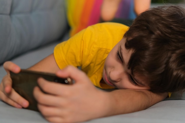 Niño soñoliento jugando en el teléfono acostado en el sofá adicción a los gadgets apatía por exceso de trabajo