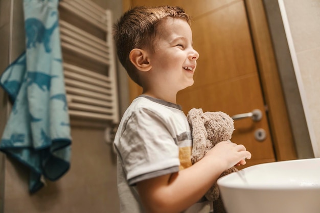 Un niño sonríe al espejo mientras está parado en el baño con su conejito de juguete por la mañana.