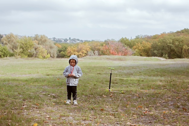 Niño sonriente de 4 años a pie con scooter en el prado verde, el niño y la naturaleza de fondo, infancia saludable activa