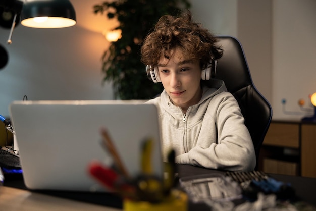 Un niño sonriente con cabello rizado que usa auriculares inalámbricos trabaja en un proyecto escolar frente a una computadora portátil hasta altas horas de la noche. Escucha música y hace su tarea.