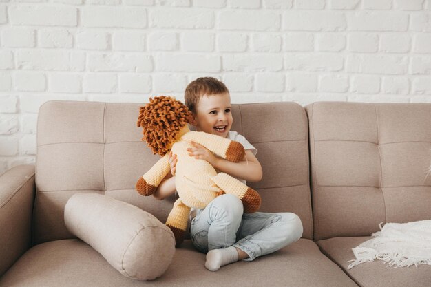 Niño sonriente con una camiseta blanca y jeans azules se sienta en el sofá y abraza a un cachorro de león de peluche