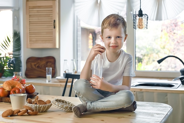 Niño sonriente caucásico sentado en la mesa de la cocina y comer galletas de avena con leche