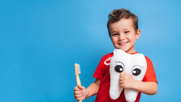 Un niño sonriente con dientes sanos sostiene un diente de felpa y un cepillo de dientes sobre un fondo azul aislado Higiene bucal Odontología pediátrica Prevención de caries Un lugar para su texto