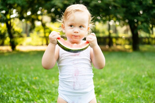 niño sonriente divertido en traje blanco comiendo sandía en el césped verde