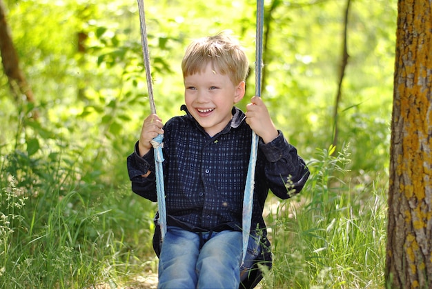 Niño sonriente divirtiéndose en el columpio de balancín tambaleante en el bosque o parque disfrutando del sol