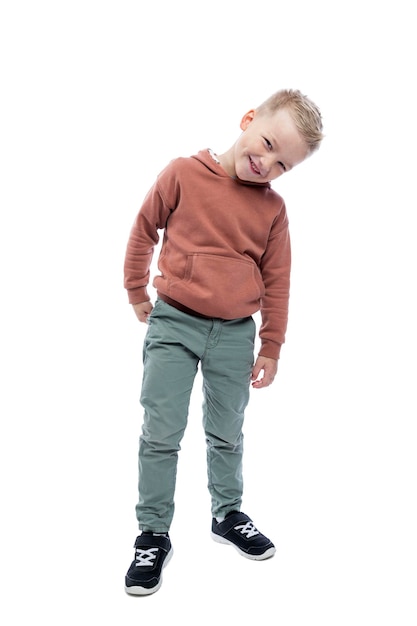 Niño sonriente está de pie Una linda rubia con una sudadera con capucha marrón y pantalones grises Altura completa Alegría y positividad Aislado sobre fondo blanco Vertical