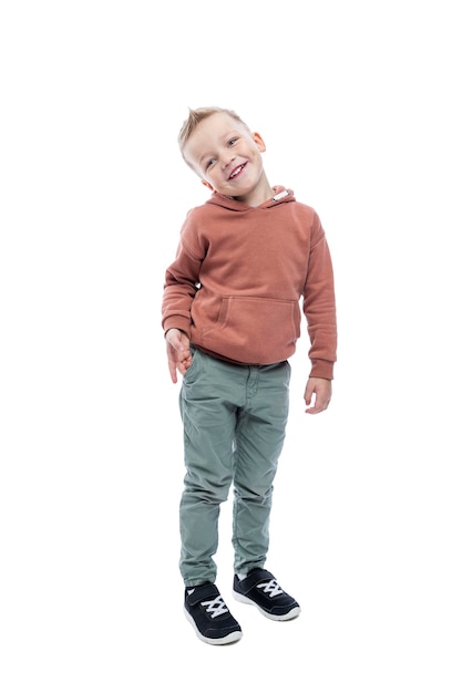 Niño sonriente está de pie Un lindo hombre rubio con un peinado de moda en una sudadera con capucha marrón y pantalones grises Alegría y positividad Altura completa Aislado sobre fondo blanco Vertical