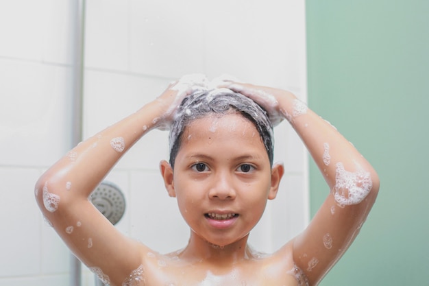 Niño sonriente frotando el cabello con champú durante la ducha en el baño.