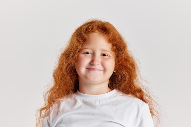 Niño sonriente linda niña pequeña con pelo rojo largo y rizado mirando a la cámara aislada sobre blanco