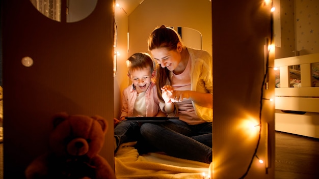 Niño sonriente con madre jugando en casa de cartón de juguete y usando tablet PC. Concepto de educación infantil y familia pasar tiempo juntos por la noche.