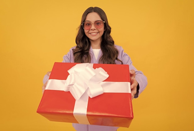 Niño sonriente de moda con gafas de sol y traje, sostenga la caja de regalo en el regalo de fondo amarillo