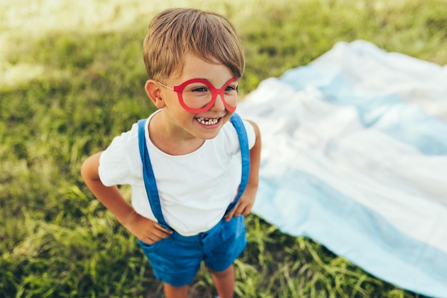 Niño sonriente con pantalones cortos azules y gafas de papel rojo jugando en el fondo de la naturaleza Niño alegre jugando en la hierba verde en el parque Niño divirtiéndose en la luz del sol al aire libre Infancia feliz