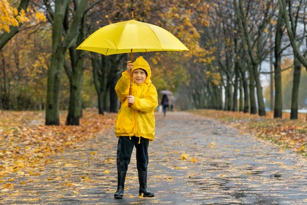 Niño sonriente en el parque con un paraguas amarillo Niño en la calle lluviosa con hojas que caen