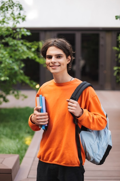 Niño sonriente de pie con mochila y libros en la mano y felizmente en el patio de la universidad