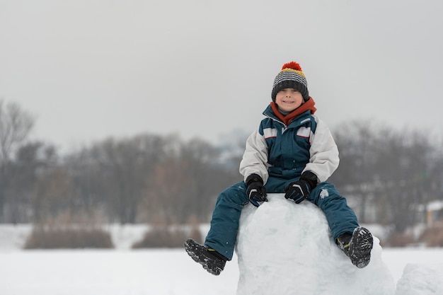 Niño sonriente de siete años se sienta en la torre de nieve. El niño construye un castillo de nieve. Horario de invierno.