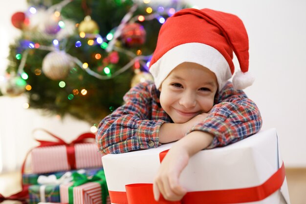 El niño de la sonrisa se sienta con una caja de regalo cerca del árbol de Navidad.