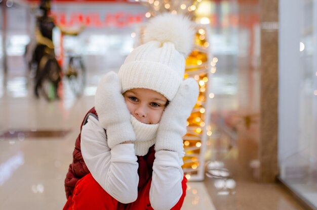 Niño sorprendido con gorro y bufanda blancos en el centro comercial cerca de las luces borrosas del escaparate