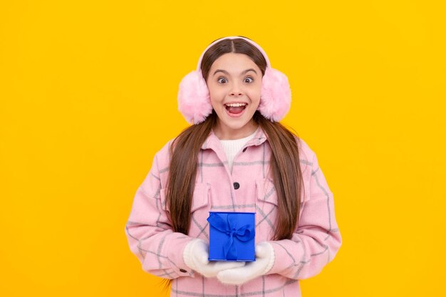 Niño sorprendido con orejeras y mitones sostiene la caja de regalo en la caja de regalo de fondo amarillo